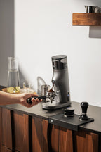Person using a Gemilai G9010C coffee grinder on a kitchen counter with a wooden shelf in the background.